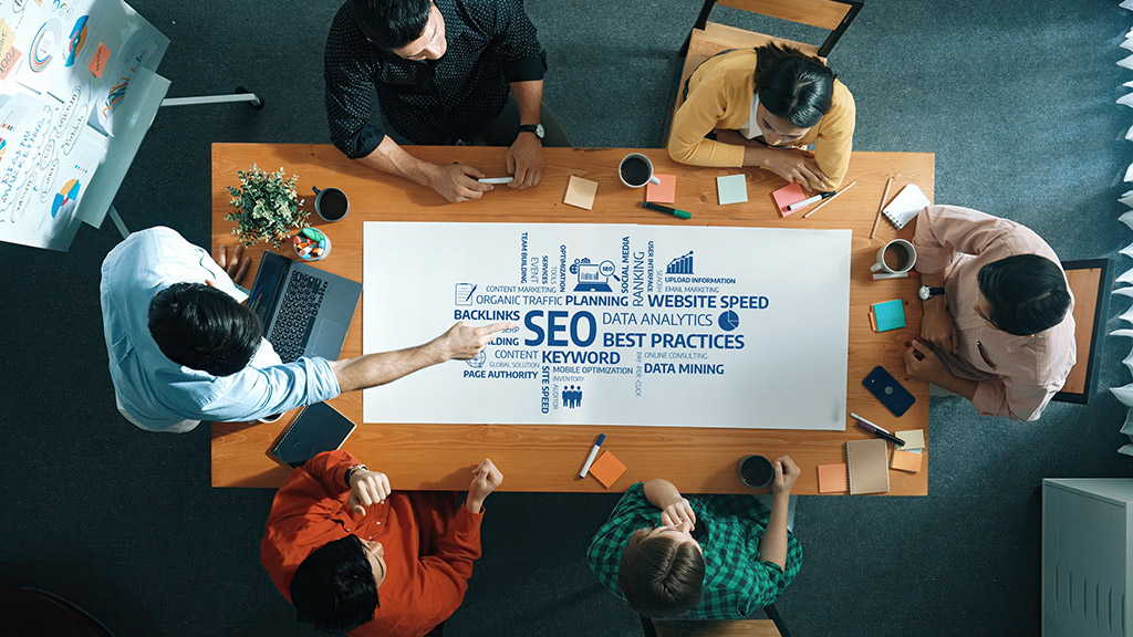 A team gathered around a table reviewing an SEO word cloud covering keywords, backlinks, website speed and data analytics.