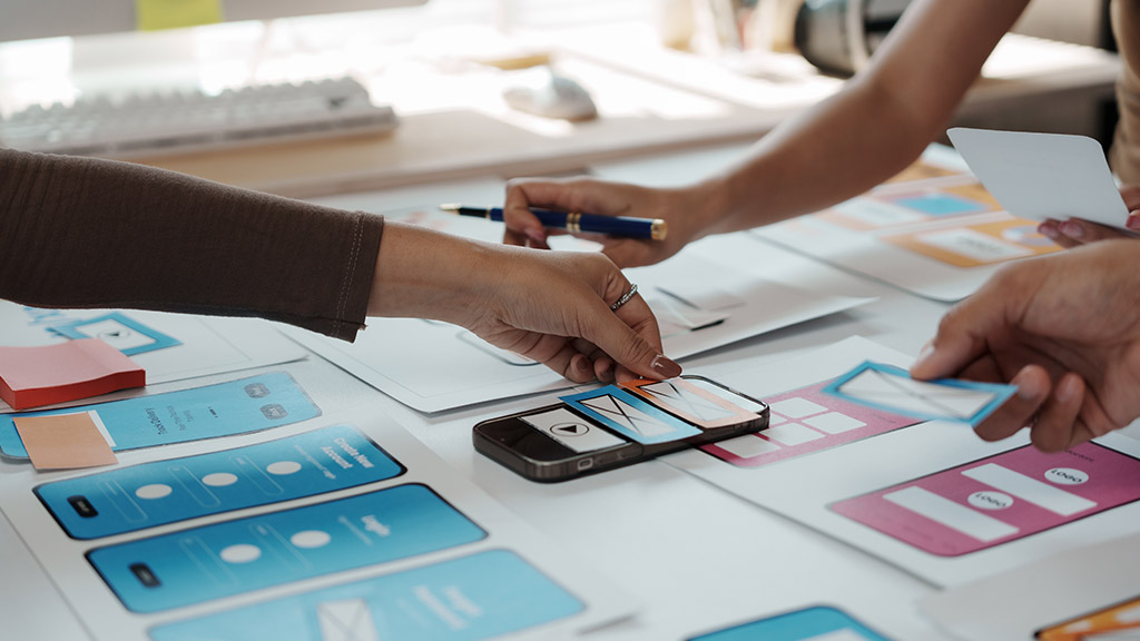 Designers arranging mobile app wireframes and a phone mockup on a table during a UX design session.
